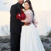 Couple posing outdoors, bride in white gown holding red bouquet beside groom in dark suit