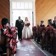 Bride and groom walking down a church aisle with guests seated on both sides, wearing red floral dresses
