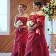 Two women in red gowns holding bouquets at an indoor wedding ceremony