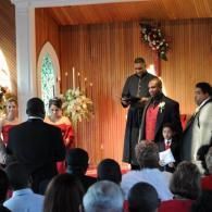Wedding ceremony inside a wood-paneled chapel with officiant, groom, bride, and guests seated.