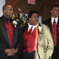 Three men in formal suits standing together indoors, wearing red vests and ties.