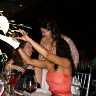 People at a dimly lit restaurant table, one woman raising her arm while smiling with wine glasses nearby