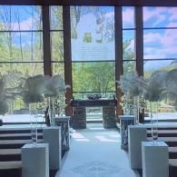 Bright church aisle with white pews, floral arrangements, and large windows overlooking greenery.