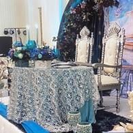 Decorated event table with blue lace cloth, floral centerpiece, and ornate white chairs under a floral arch