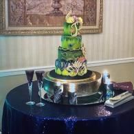 Green and black tiered cake on a silver stand at a decorated table with champagne flutes