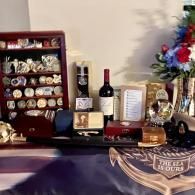 Memorial display with wine bottles, framed photos, plaques, flowers, and a folded flag on a table