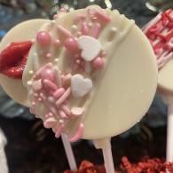 White chocolate lollipop decorated with pink sprinkles and heart candies on a stick