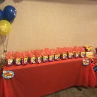 Red table with rows of small colorful party favor bags and blue-yellow balloons at the left.