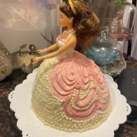 Doll-shaped cake with pink and cream ruffled icing on a white scalloped stand