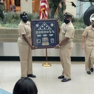 Two uniformed officers hold a framed display during a formal ceremony indoors.
