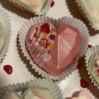 Pink heart-shaped dessert decorated with sprinkles in a white paper cup, surrounded by similar sweets
