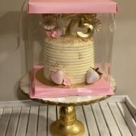 Pink and white cake in a clear display box on a gold stand, decorated with a gold number 3 and hearts