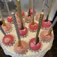 Pink cake pops with gold sticks on a white frosted cake stand, decorated with sprinkles.