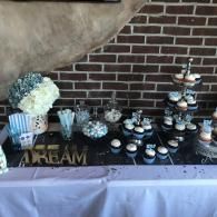 Dessert table with blue and white cupcakes, cake pops, and a white floral arrangement against a brick wall