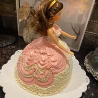 Pink princess doll cake with gold accents on a white scalloped base, shown on a countertop
