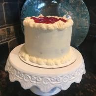 White frosted cake with red topping on a decorative white stand, set against a blue glass backdrop