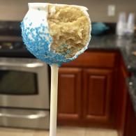 Partially eaten blue-and-white cake pop on a stick in a kitchen, with a blurred stove and cabinets behind it
