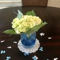 Blue vase of pale yellow flowers on a lace doily, with scattered white and blue confetti on a dark table.