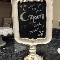 Decorative chalkboard sign on a white stand with “Love you to the moon and back” and star doodles.
