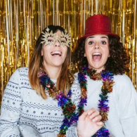 Two smiling people in festive hats and tinsel posing before a gold streamers backdrop