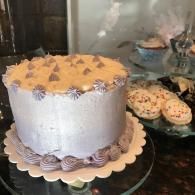 Tall lavender frosted cake with cookie topping on a stand, beside decorated cupcakes on a dark counter