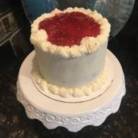 White cake with whipped cream border and red fruit topping on a decorative cake stand