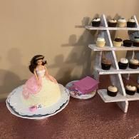 Mini doll in pink dress on a plate beside a small dessert display stand with pastries.