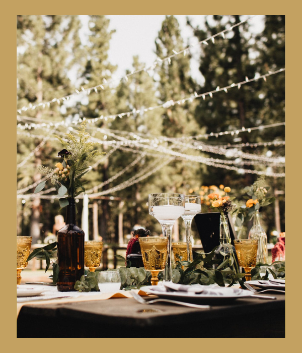 Outdoor banquet table with glassware and flowers under white string lights in a garden setting