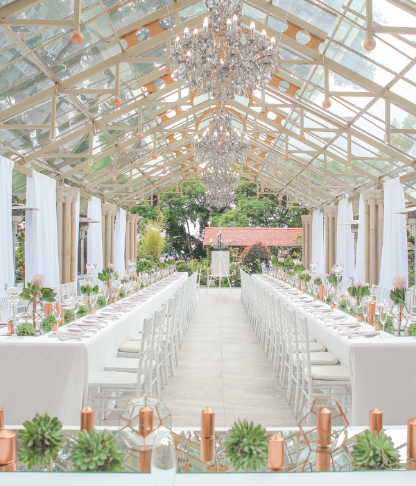 Elegant glass-roofed banquet hall with long white tables, chandeliers, and floral decor