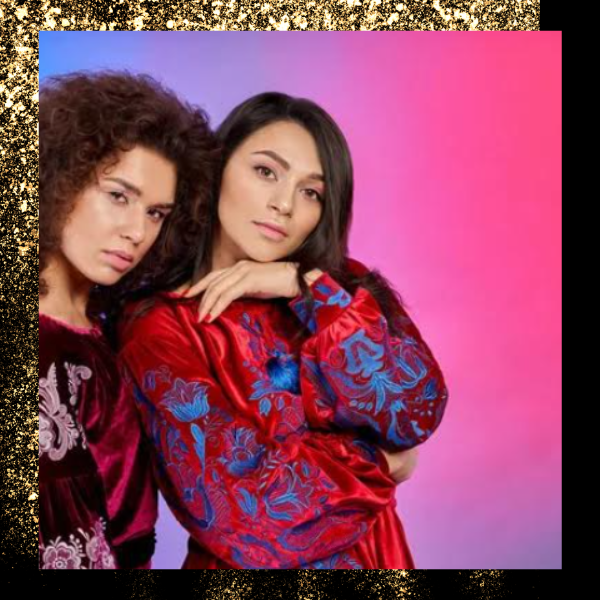 Two women posing against a pink gradient backdrop, wearing vibrant red and purple patterned outfits.