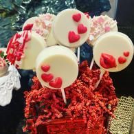 Heart-shaped cake pops in white chocolate with red hearts, nestled in red crinkle paper.