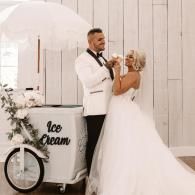 Bride and groom serving ice cream from a white cart at a wedding reception