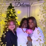 Three people smiling at a holiday event, standing by a lit Christmas tree and “Delores” backdrop.