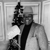 Smiling person in a hat holding a “YOLO” sign in front of a Christmas tree