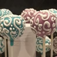Teal and purple frosted cake pops with white swirls on sticks arranged in a row