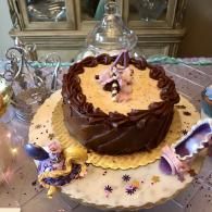 Two-tier chocolate-frosted cake with purple and gold decorations on a white tablecloth