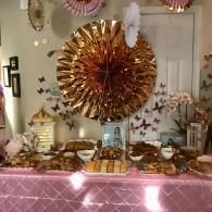 Pink and gold dessert table with butterfly decor and a large rosette backdrop in a home setting