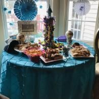 Table with a teal cloth at a decorated dessert buffet, featuring cakes, treats, and blue party decorations.