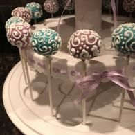 Decorated cake pops on sticks arranged on a white stand with a purple ribbon.