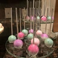 Pastel pink and mint cake pops on sticks in a clear display stand on a table.