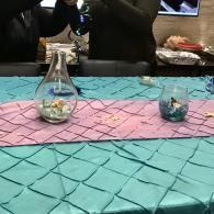 Two glass vases on a turquoise and pink table runner, with people blurred in the background.