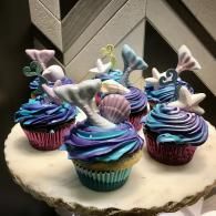 Six pastel frosted cupcakes on a round cake board against a chevron backdrop.