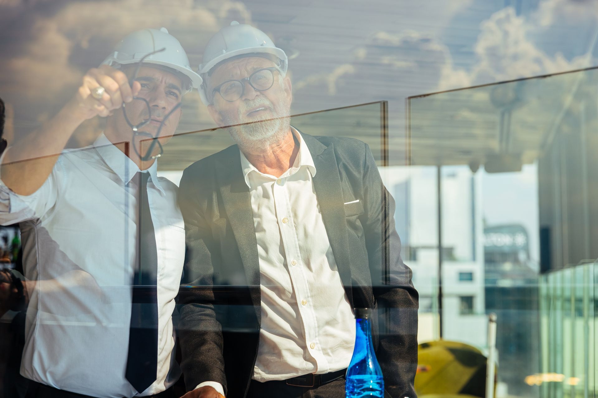 Two men wearing hard hats and glasses are standing next to each other in front of a window.