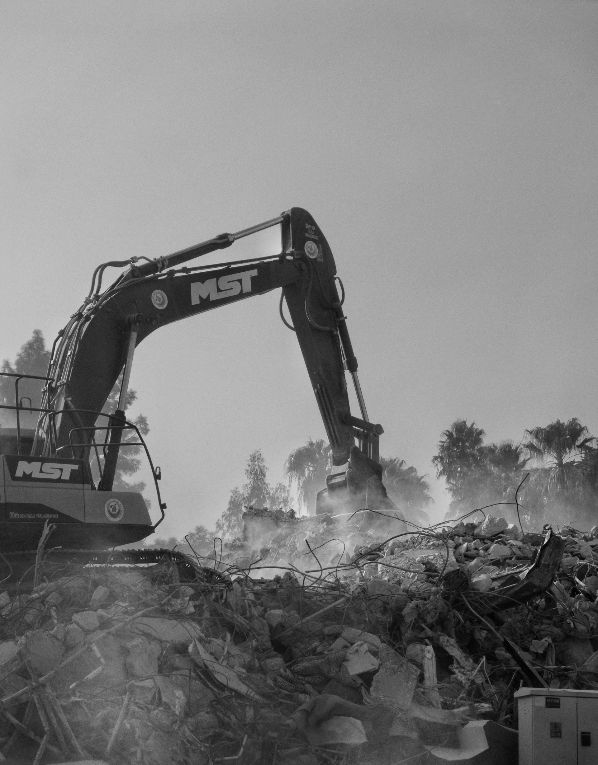 Excavator demolishing a building, kicking up dust; bright sky in the background.