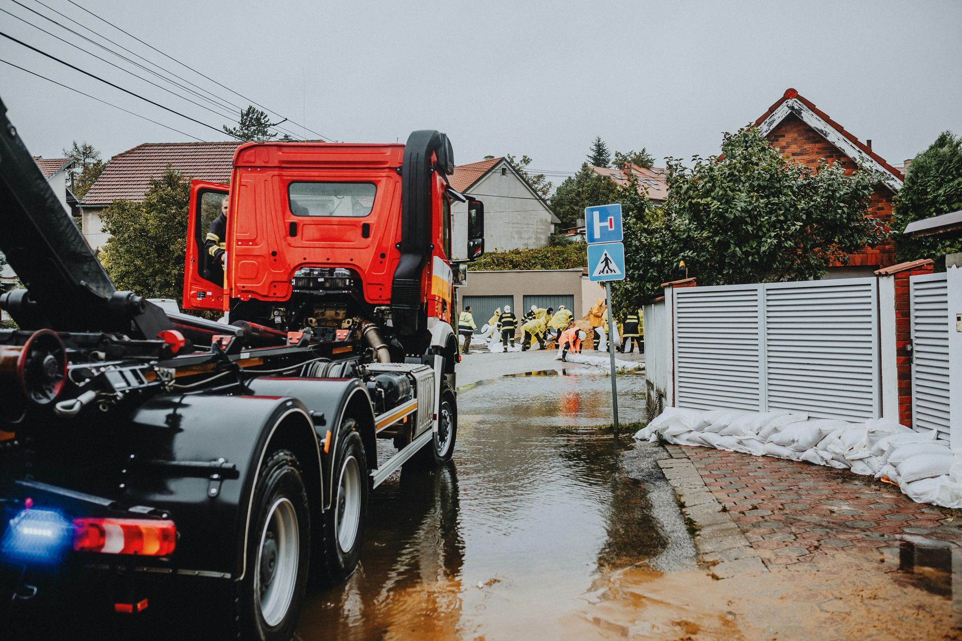 Fire truck in floodwater, rescue workers in background, sandbags along a white fence.