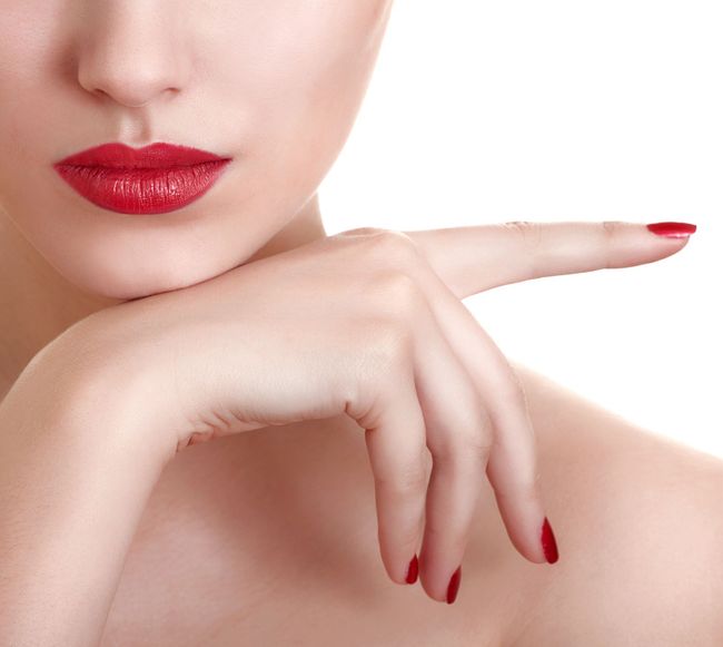 Primer plano de labios rojos y mano con manicura y uñas rojas apuntando hacia un lado sobre un fondo blanco.