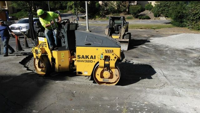 Large circular pothole in a gray asphalt road near the curb, with cracked pavement around it.