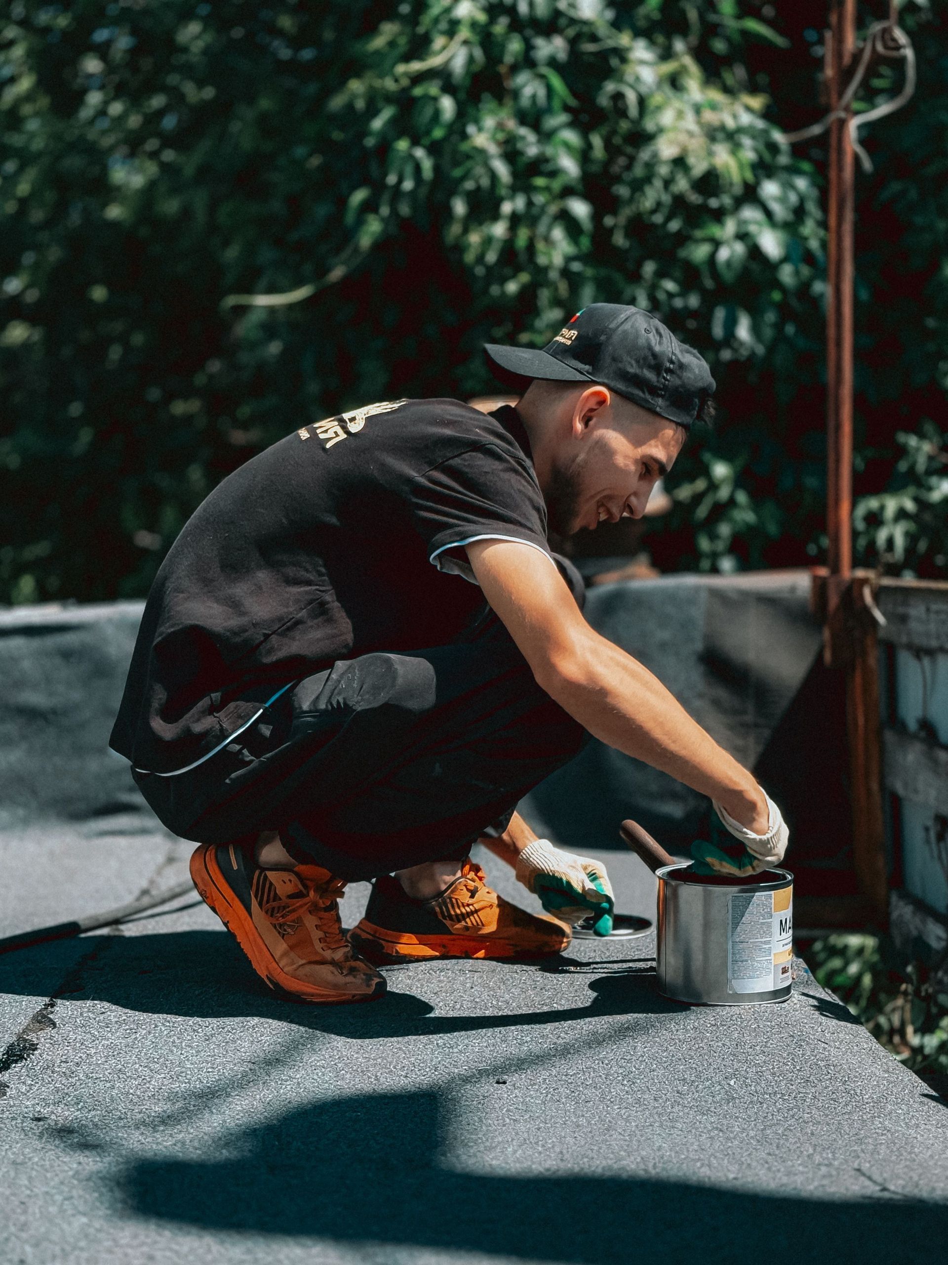 Man kneeling outdoors, painting a surface with a brush and paint can beside him