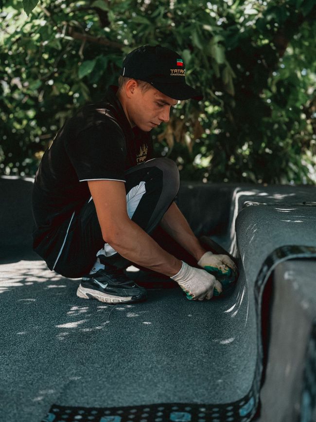 Person crouching on a truck bed, fixing or cleaning the surface with a tool, outdoors in shade.