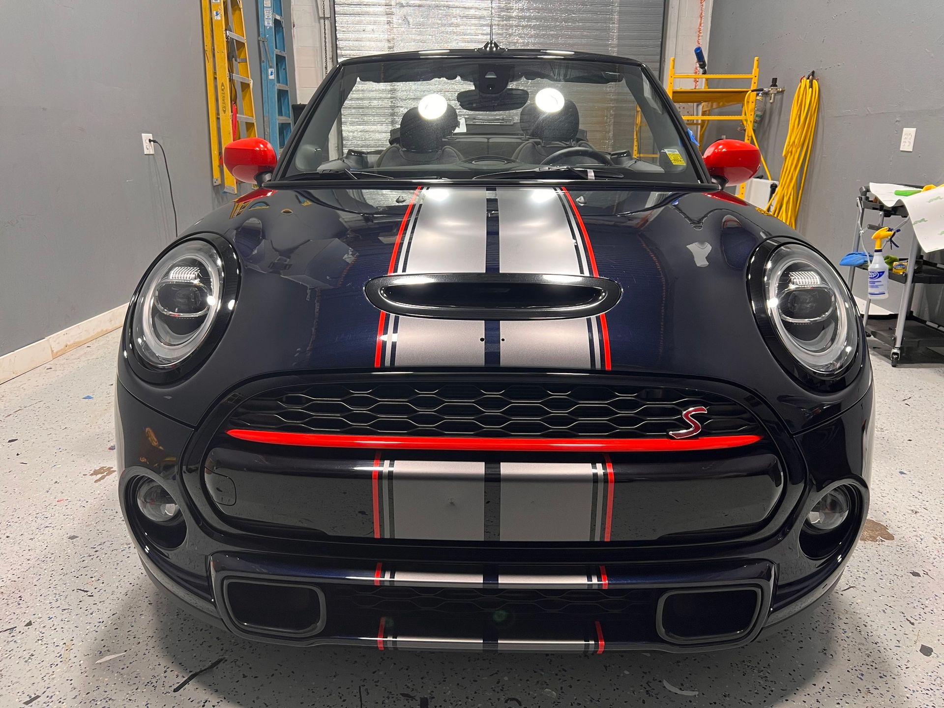 Front view of a black Mini Cooper with white racing stripes in a garage.